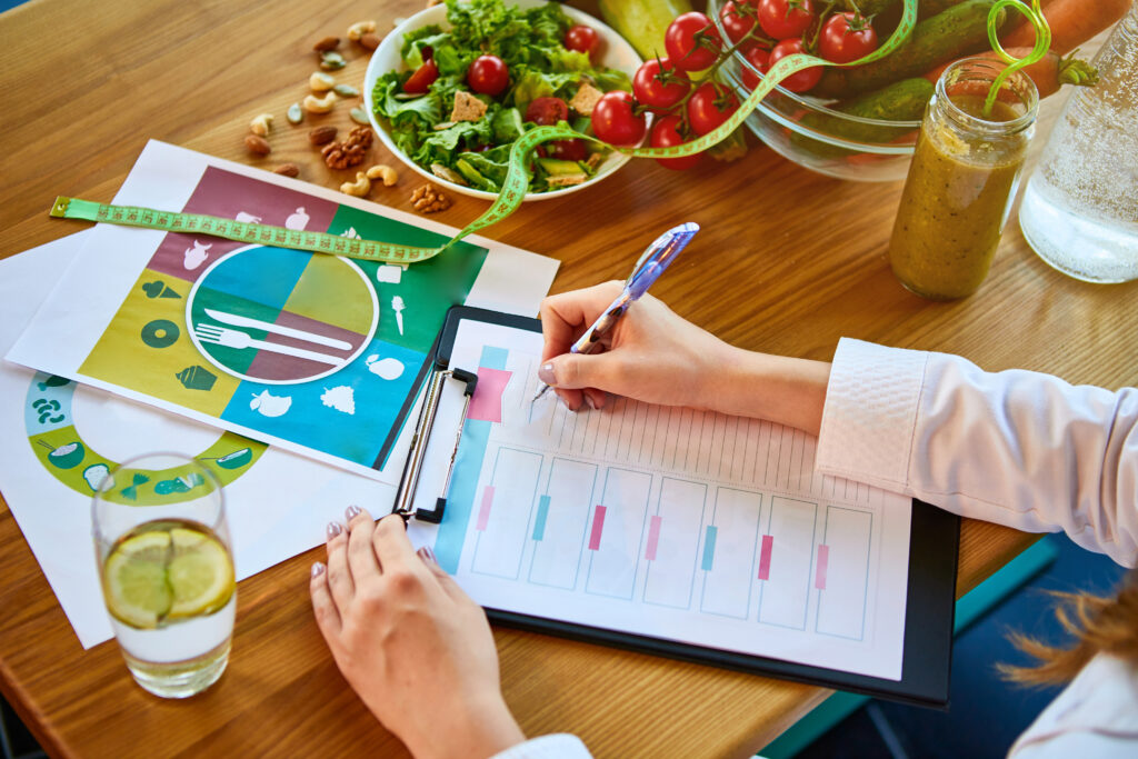 Woman dietitian in medical uniform with tape measure working on a diet plan sitting with different healthy food ingredients in the green office on background. Weight loss and right nutrition concept